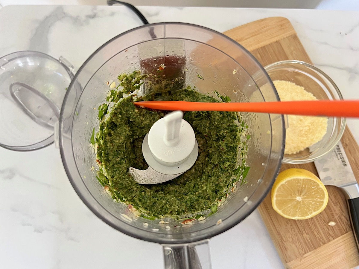 Orange spatula scraping down the sides of the food processor after blending pesto for Pesto Pasta with Tomatoes.