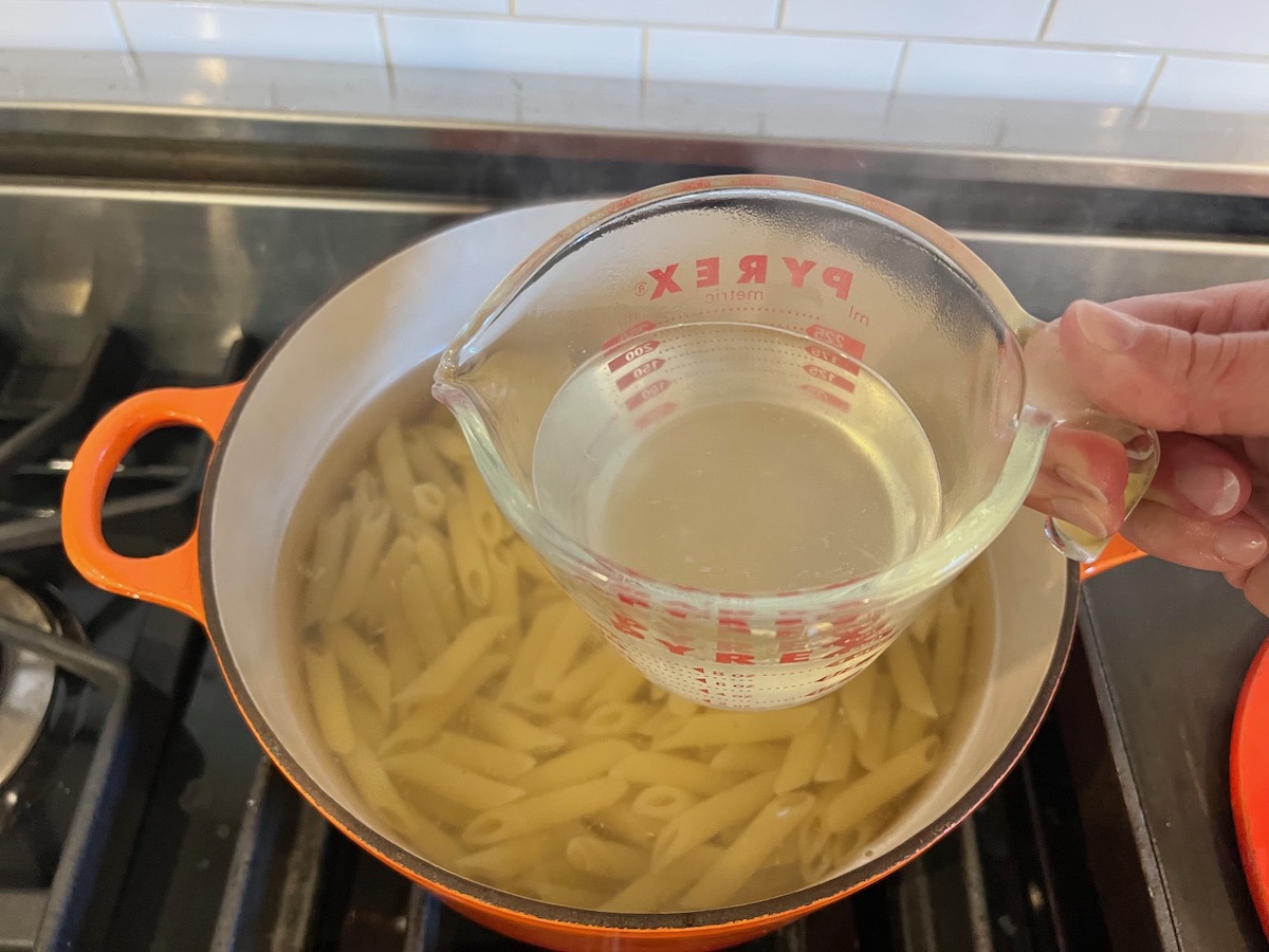 Measuring cup with pasta water over pot with pasta cooking for Pesto Pasta with Tomatoes.