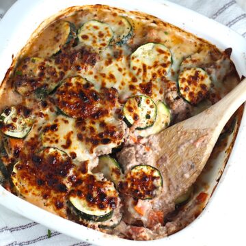 Ground Beef and Zucchini Casserole baked golden brown on top with wood spatula in corner of dish after some is scooped out. Sitting on blue and white striped dish towel on counter.