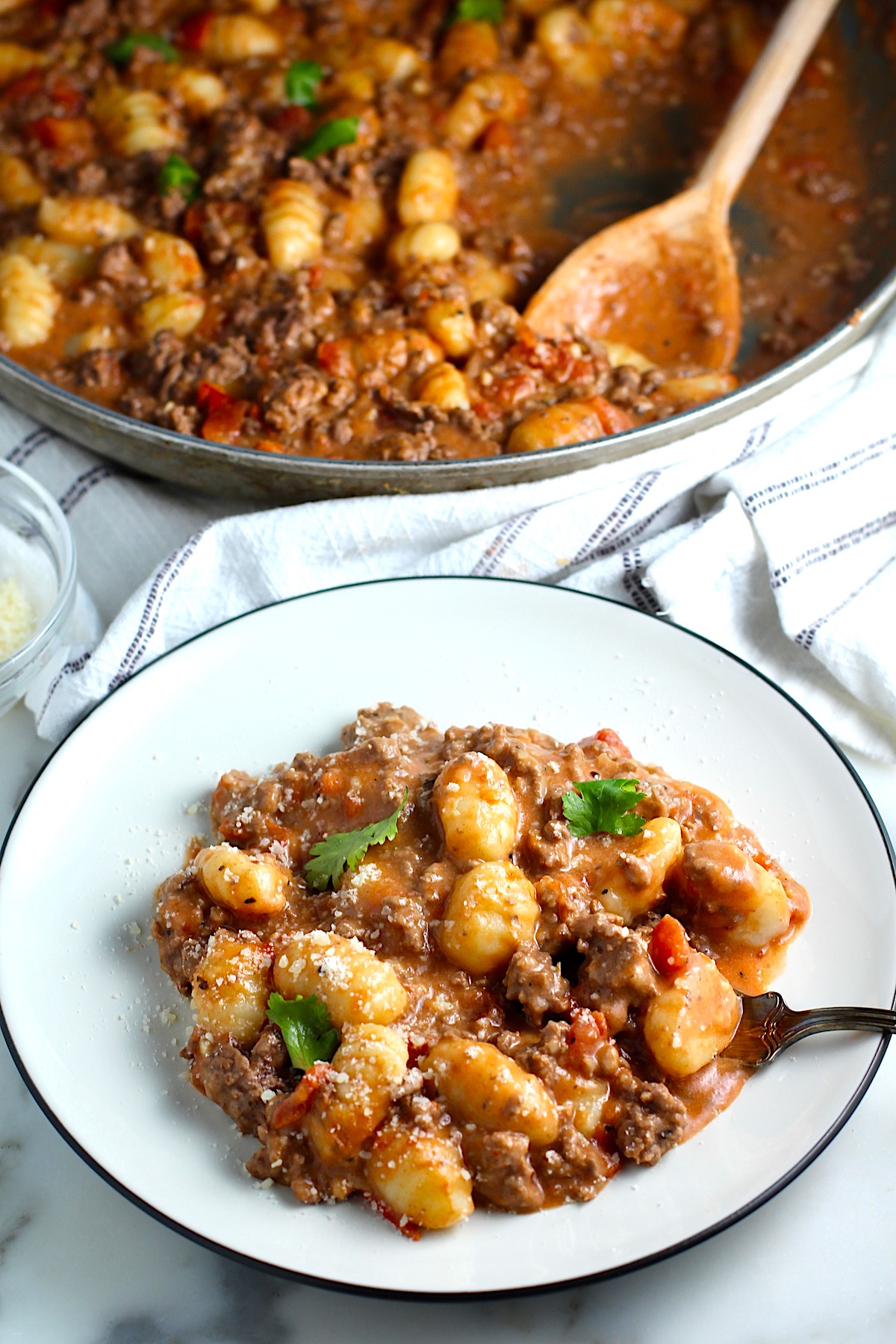 Ground Beef Gnocchi Skillet recipe on a plate with fresh parsley garnish and a fork. In the background is the skillet with the Beef Gnocchi and a wood spatula.
