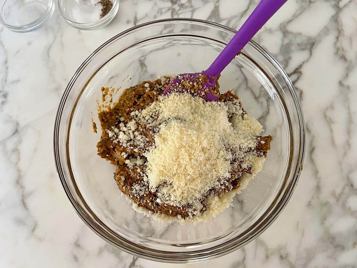 Grated parmesan cheese added to a bowl of Sun Dried Tomato Pesto with a spatula in the bowl ready to mix to combine.