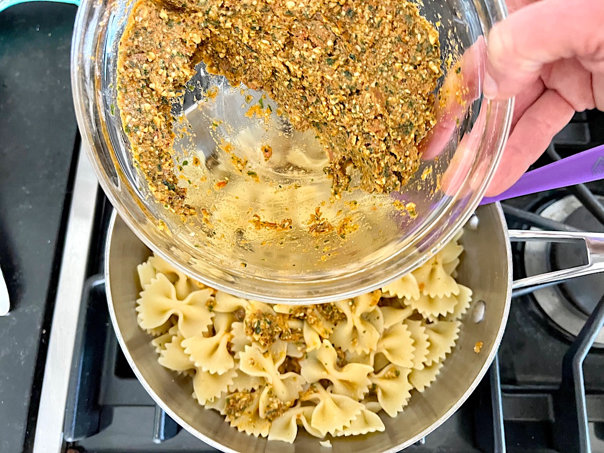 Hand holding a bowl of Sun Dried Tomato Pesto in a clear glass bowl over a pot of drained bow tie pasta as the pesto is added to the pasta.