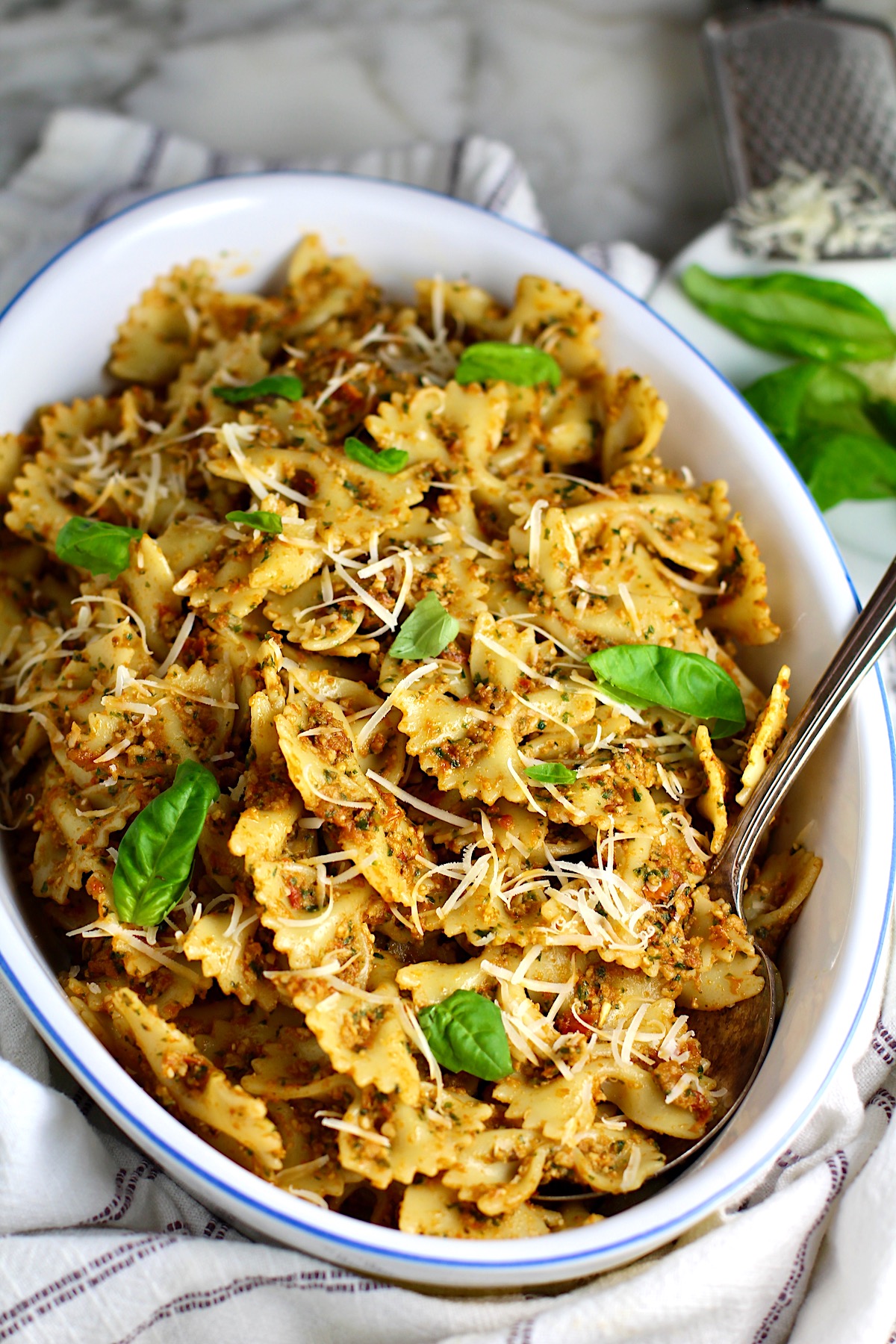 Sun Dried Tomato Pesto Pasta in an oval serving platter with serving spoon on a blue and white striped towel on the counter.  Basil leaves in the upper right corner of the picture on the counter.