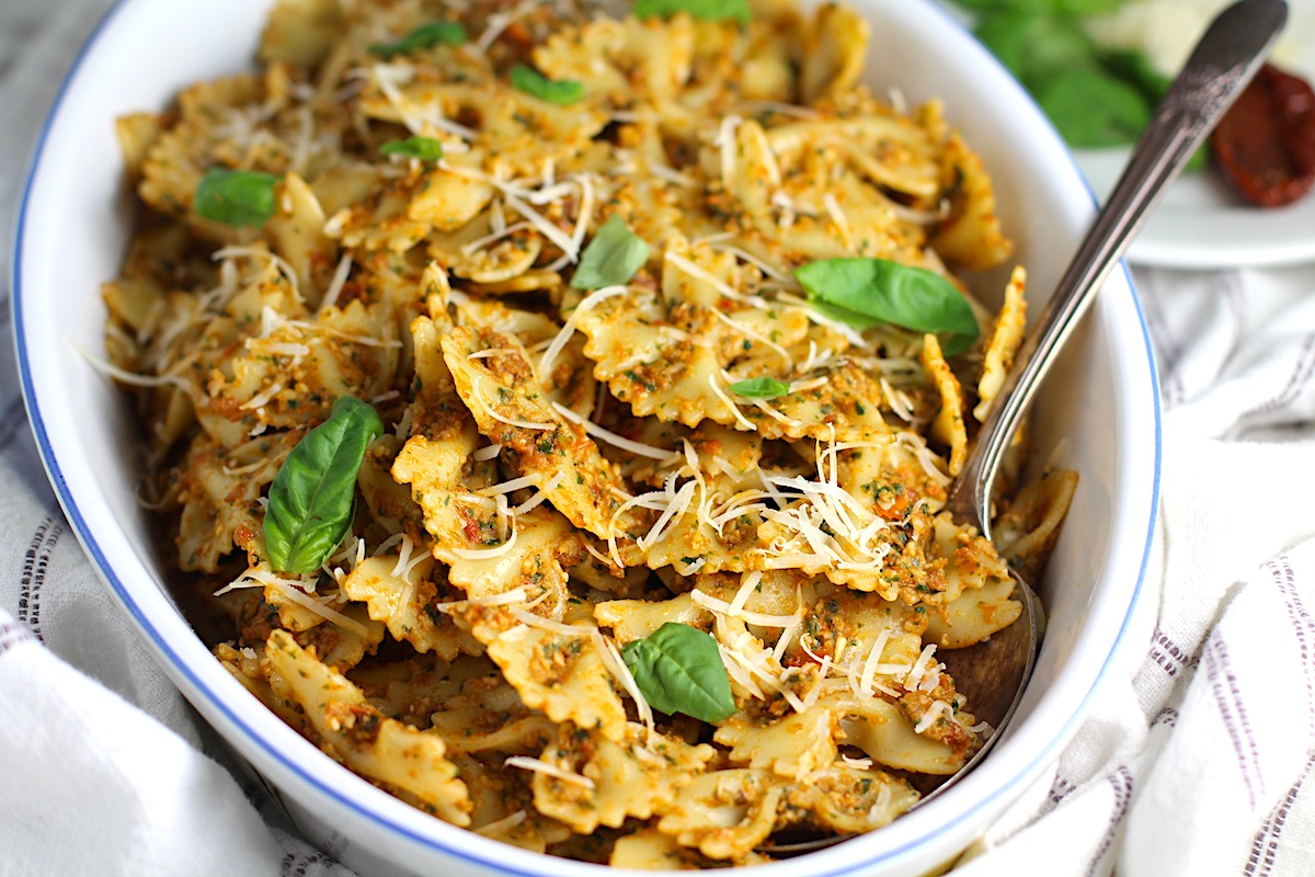 Sun Dried Tomato Pesto Pasta in an oval serving platter with serving spoon on a blue and white striped towel on the counter. Basil leaves in the upper right corner of the picture on the counter.