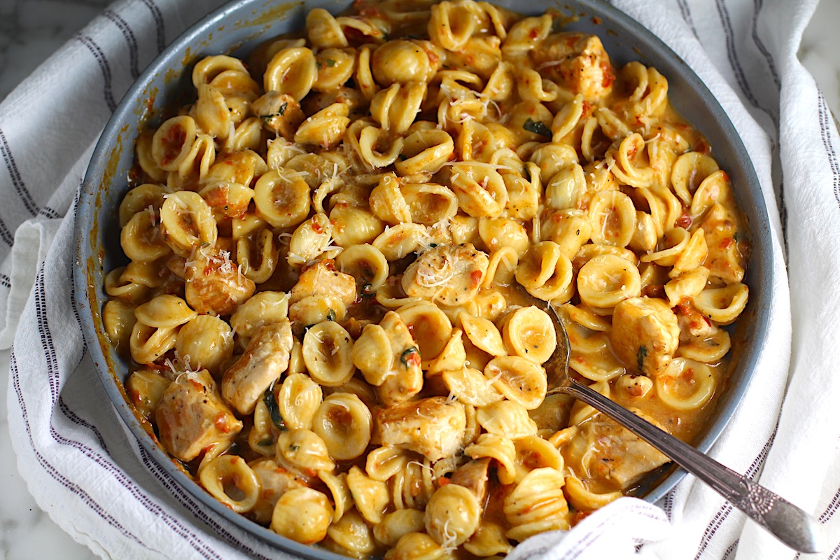 Creamy Sun Dried Tomato Sauce with Chicken and Pasta with serving spoon in a large pan on a kitchen towel that is white with blue stripes.