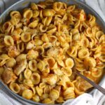 Creamy Sun Dried Tomato Sauce with Chicken and Pasta with serving spoon in a large pan on a kitchen towel that is white with blue stripes.