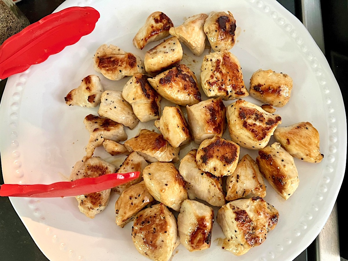 Browned chicken cubes being transferred to a white plate with red silicone tongs for Creamy Sun Dried Tomato Sauce with Chicken and Pasta recipe.