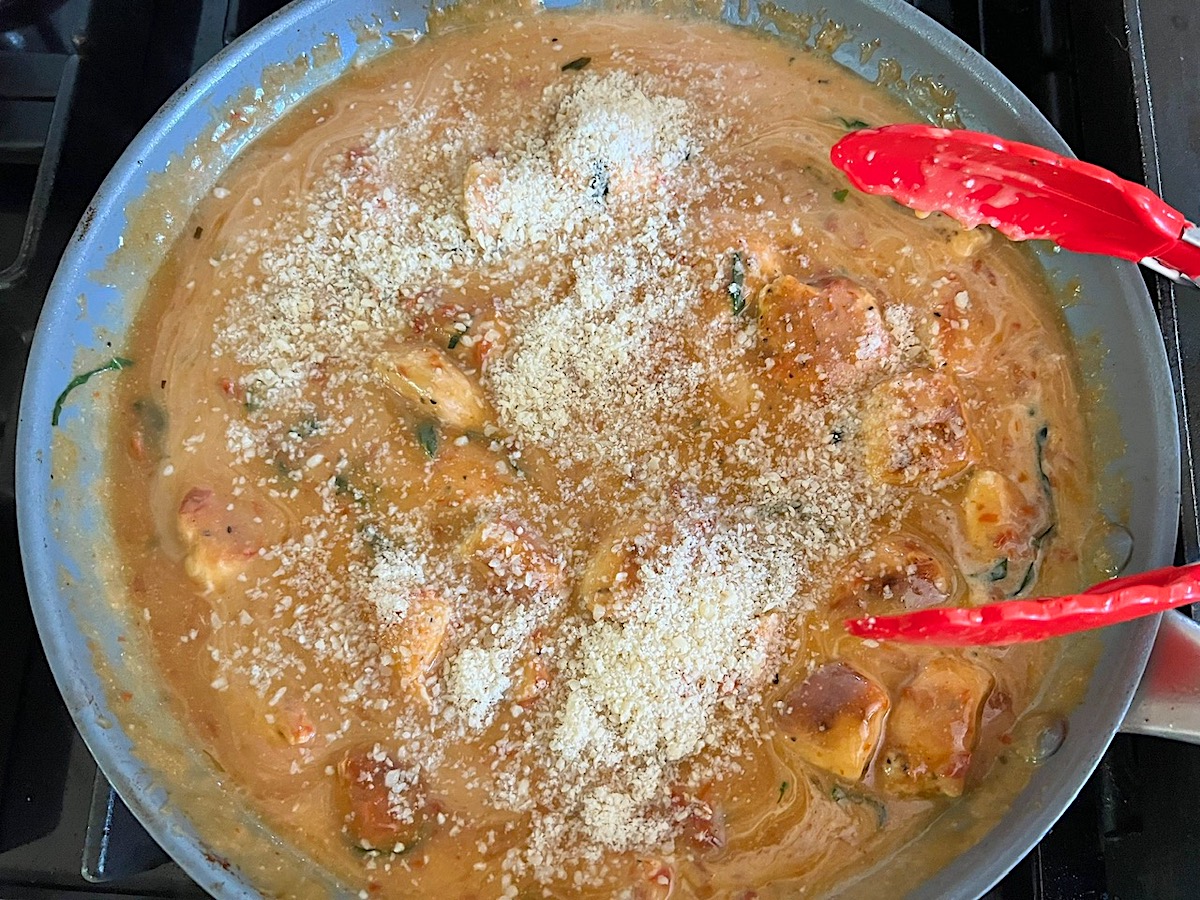 Cooked chicken and grated parmesan cheese added to the thickened sun dried tomato sauce in a large pan on stove for the Creamy Sun Dried Tomato Sauce with Chicken and Pasta recipe.