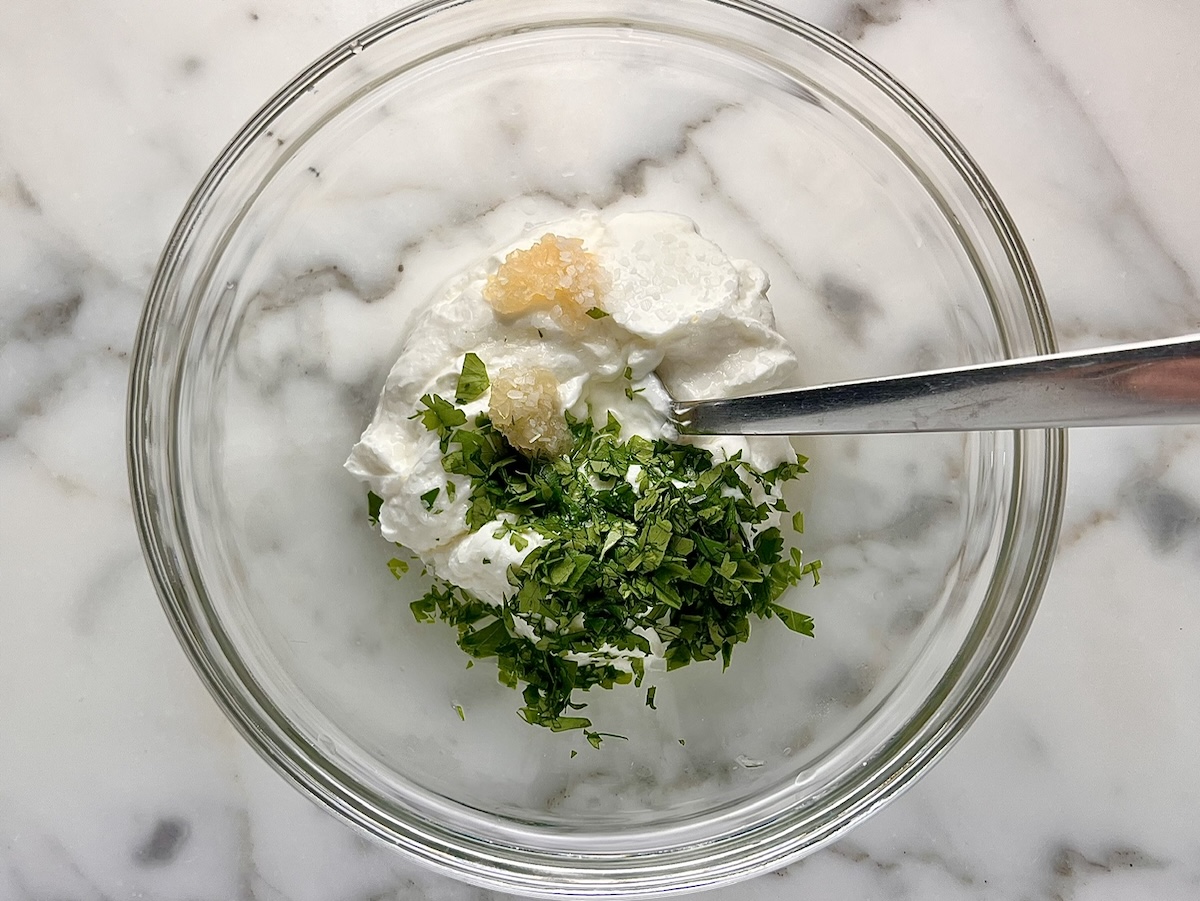 Yogurt, minced garlic, chopped cilantro, and lemon juice in a clear glass bowl before being mixed by fork to become the Cilantro Yogurt sauce to go in the Chicken Tikka Wraps.