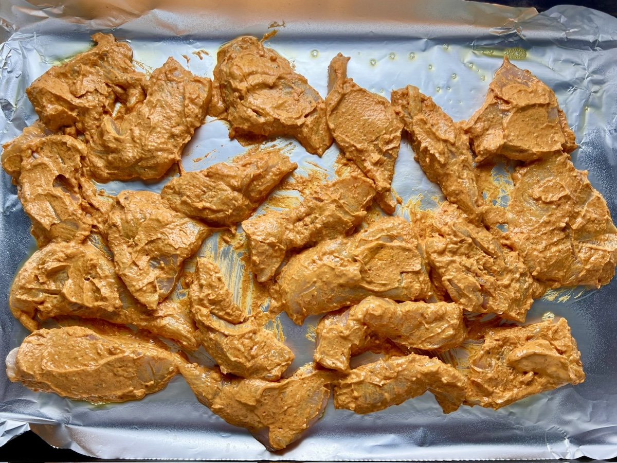 Raw chicken strips coated in tikka yogurt marinade spread out on a foil-lined sheet pan before being roasted for the Chicken Tikka Wraps.