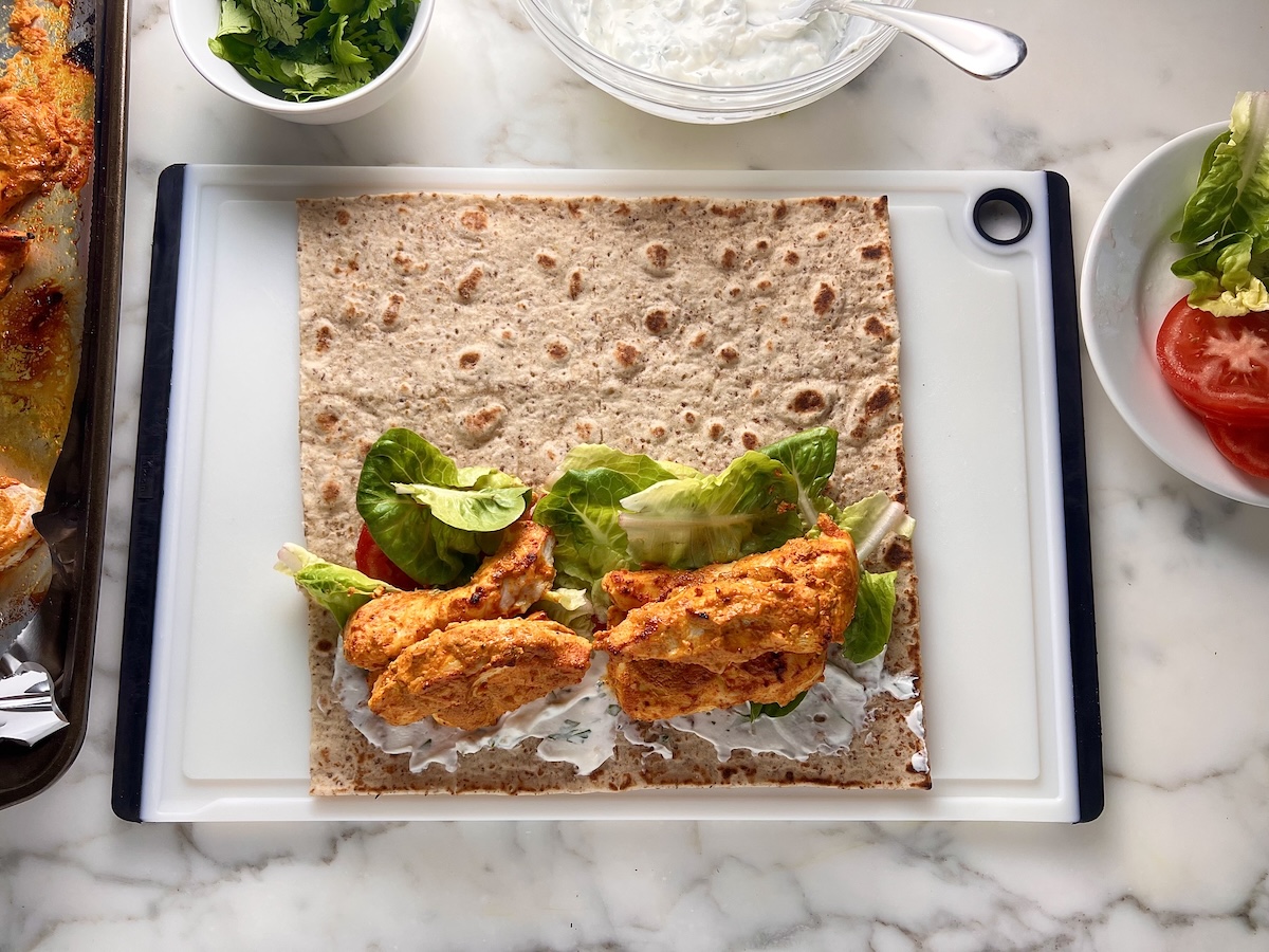 Large square flatbread wrap laying on a white cutting board on the counter with bowls of cilantro, yogurt sauce, and the pan of cooked chicken around for assembling the Chicken Tikka Wraps.
