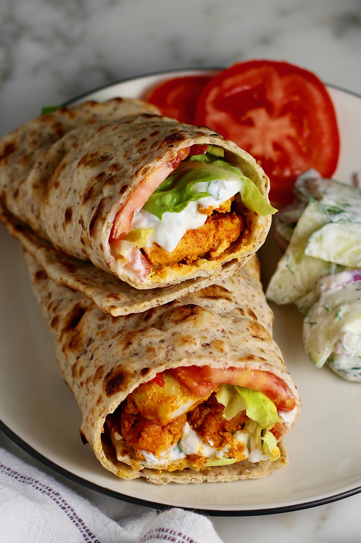 Two Chicken Tikka Wraps stuffed with seasoned chicken, lettuce, tomato, and yogurt sauce stacked on top of each other on a plate with a creamy cucumber salad on the side and two slices of tomato. The plate is on a marble counter with a white towel with blue stripes.