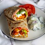 Two Chicken Tikka Wraps stuffed with seasoned chicken, lettuce, tomato, and yogurt sauce stacked on top of each other on a plate with a creamy cucumber salad on the side and two slices of tomato. The plate is on a marble counter with a white towel with blue stripes.