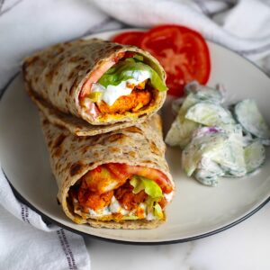 Two Chicken Tikka Wraps stuffed with seasoned chicken, lettuce, tomato, and yogurt sauce stacked on top of each other on a plate with a creamy cucumber salad on the side and two slices of tomato. The plate is on a marble counter with a white towel with blue stripes.