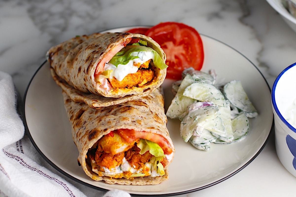 Two Chicken Tikka Wraps stuffed with seasoned chicken, lettuce, tomato, and yogurt sauce stacked on top of each other on a plate with a creamy cucumber salad on the side and two slices of tomato. The plate is on a marble counter with a white towel with blue stripes.