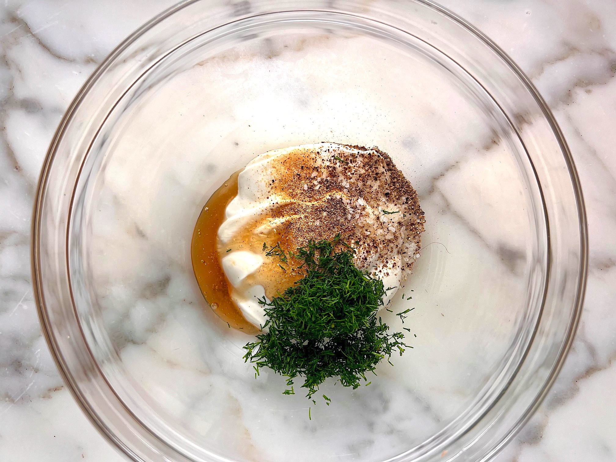 Sour cream, vinegar, honey, fresh dill, salt, and pepper, all added to the bottom of a clear bowl on the counter before being mixed for the Dill and cucumber salad recipe.