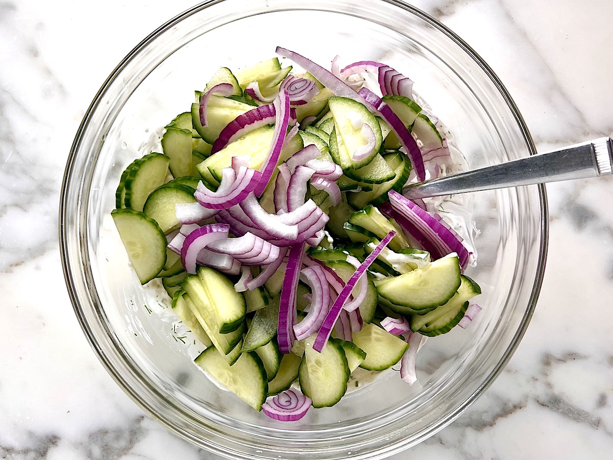 Sliced half moon-shaped cucumber with the skin on and thinly sliced red onion added to the clear bowl on the counter before being mixed for the Dill and cucumber salad recipe.
