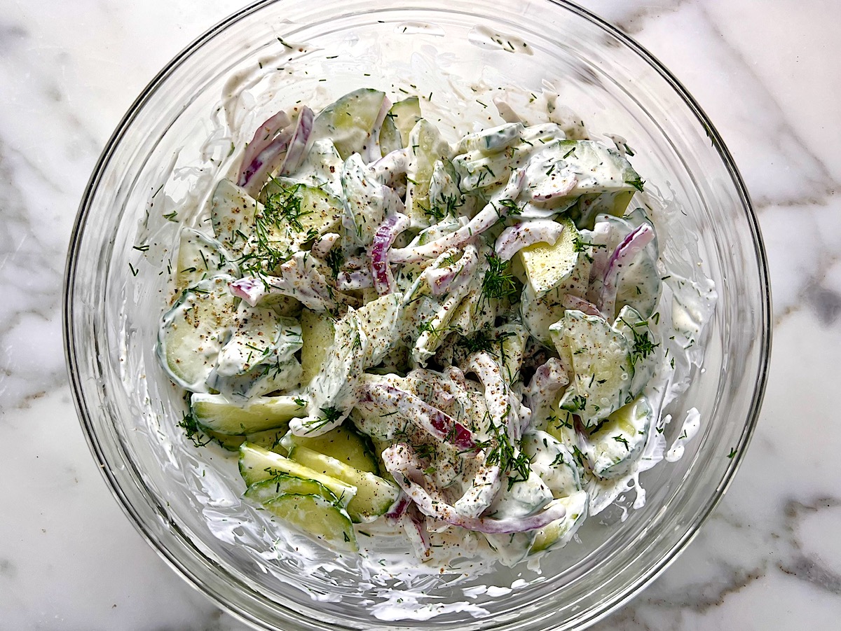 Creamy Dill and Cucumber Salad with thinly sliced cucumber and red onion in a sour cream dressing mixed in a clear mixing bowl on the counter.