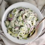 Creamy Dill and Cucumber Salad with thinly sliced cucumber and red onion in a sour cream dressing in a bowl with a serving spoon on the counter next to a blue and white striped towel.