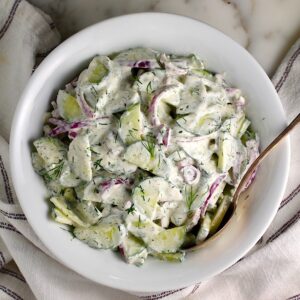 Creamy Dill and Cucumber Salad with thinly sliced cucumber and red onion in a sour cream dressing in a bowl with a serving spoon on the counter next to a blue and white striped towel.
