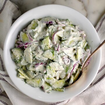 Creamy Dill and Cucumber Salad with thinly sliced cucumber and red onion in a sour cream dressing in a bowl with a serving spoon on the counter next to a blue and white striped towel.