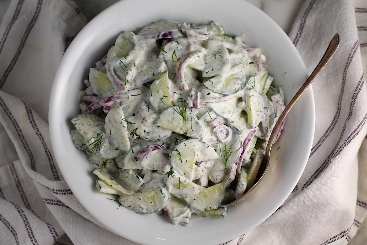 Creamy Dill and Cucumber Salad with thinly sliced cucumber and red onion in a sour cream dressing in a bowl with a serving spoon on the counter next to a blue and white striped towel.