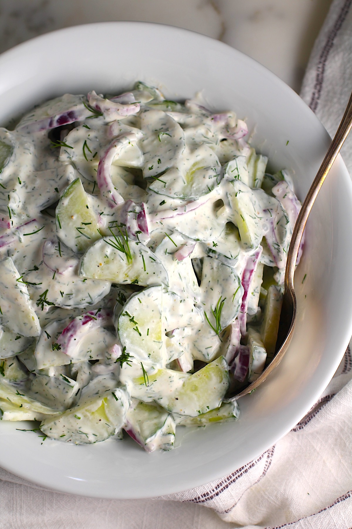 Creamy Dill and Cucumber Salad with thinly sliced cucumber and red onion in a sour cream dressing in a bowl with a serving spoon on the counter next to a blue and white striped towel.