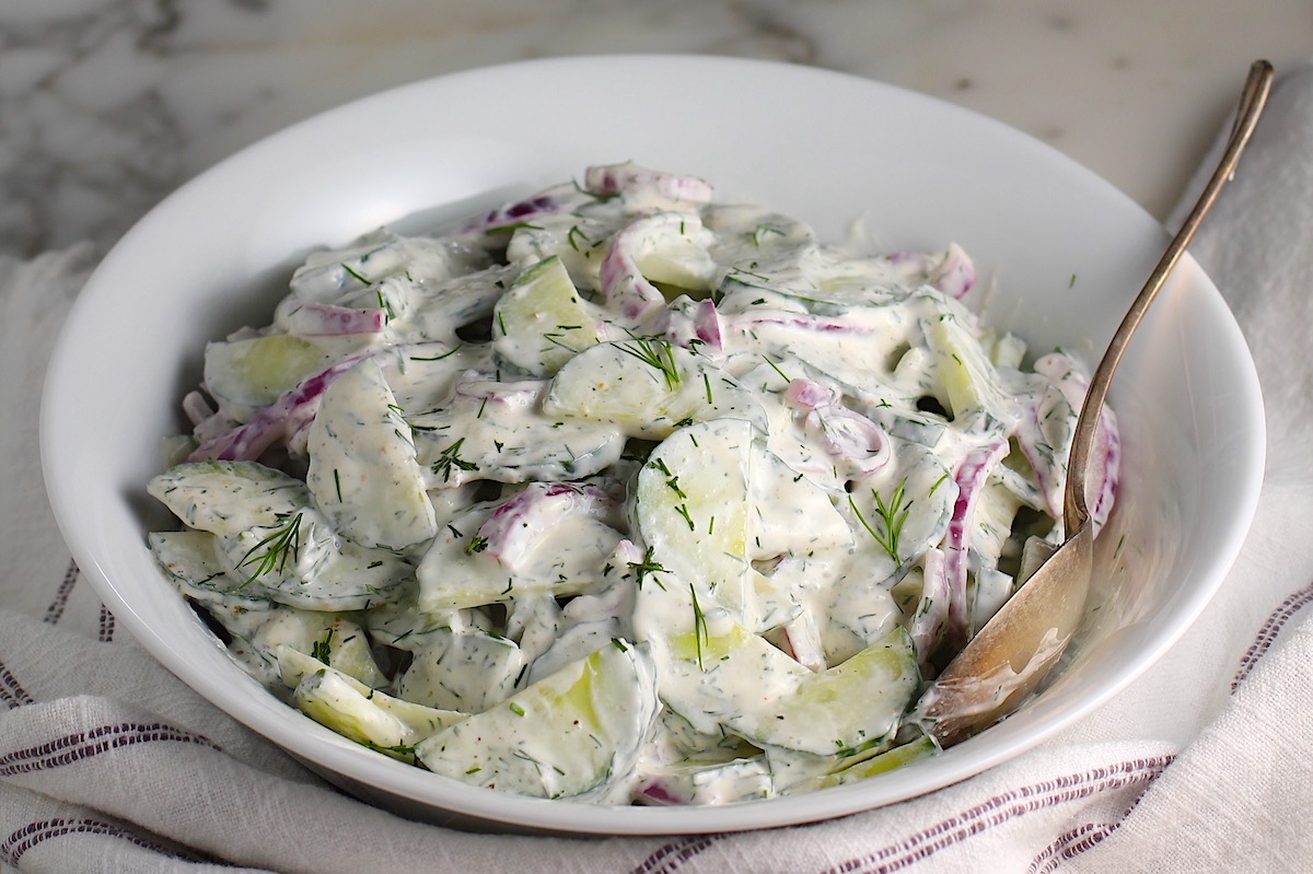 Creamy Dill and Cucumber Salad with thinly sliced cucumber and red onion in a sour cream dressing in a bowl with a serving spoon on the counter next to a blue and white striped towel.