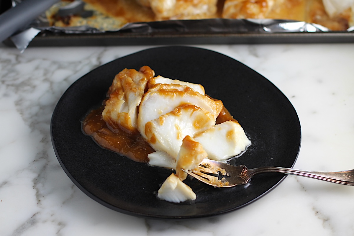 One filet of cooked miso glazed cod that has been broken into pieces on a black plate and a fork sitting on the plate with piece of cod on it. There is a sheet pan with the other filets in background for Glazed Miso Cod Recipe.