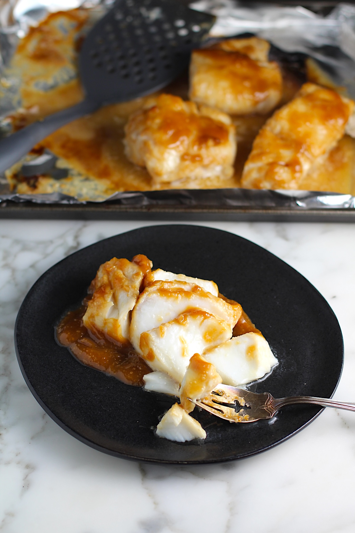 One filet of cooked miso glazed cod that has been broken into pieces on a black plate and a fork sitting on the plate with piece of cod on it. There is a sheet pan with the other filets in background for Glazed Miso Cod Recipe.