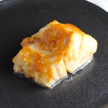 One filet of cooked miso glazed cod on a black plate with golden brown miso sauce on top of it for the Glazed Miso Cod Recipe.