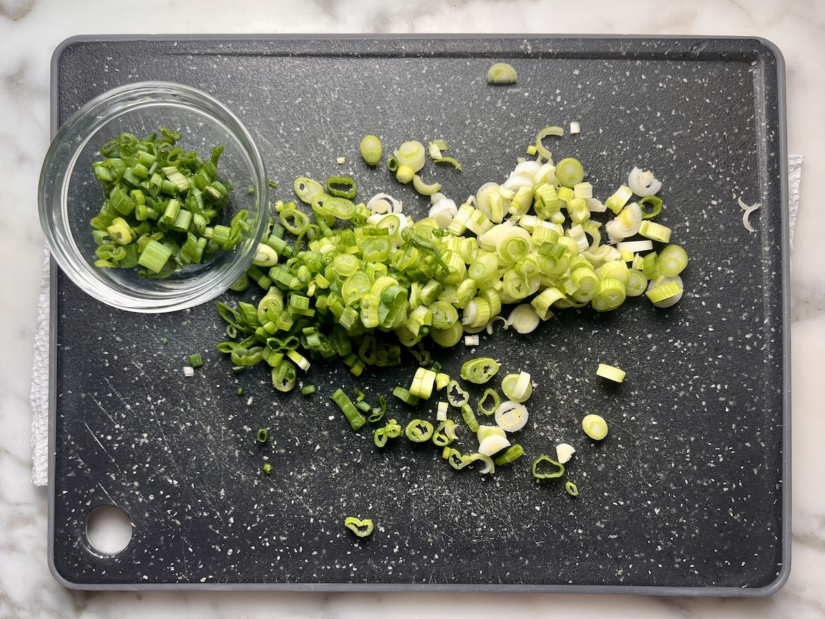 Sliced scallions on black cutting board with some of the greens in a clear bowl to be used for garnish for Stuffed Peppers with Ground Turkey recipe.