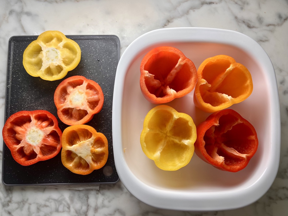 Four red, orange, and yellow bell peppers hollowed out and sitting in a casserole dish with the tops sitting next to it on the cutting board for Stuffed Peppers with Ground Turkey recipe.