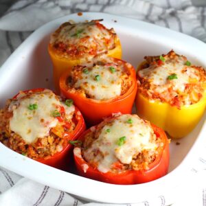 Five Red and Yellow Stuffed peppers with Ground Turkey, rice, and topped with melted cheese and a sprinkle of sliced scallions. The peppers are in the large square casserole dish on a blue and white striped towel on the counter.