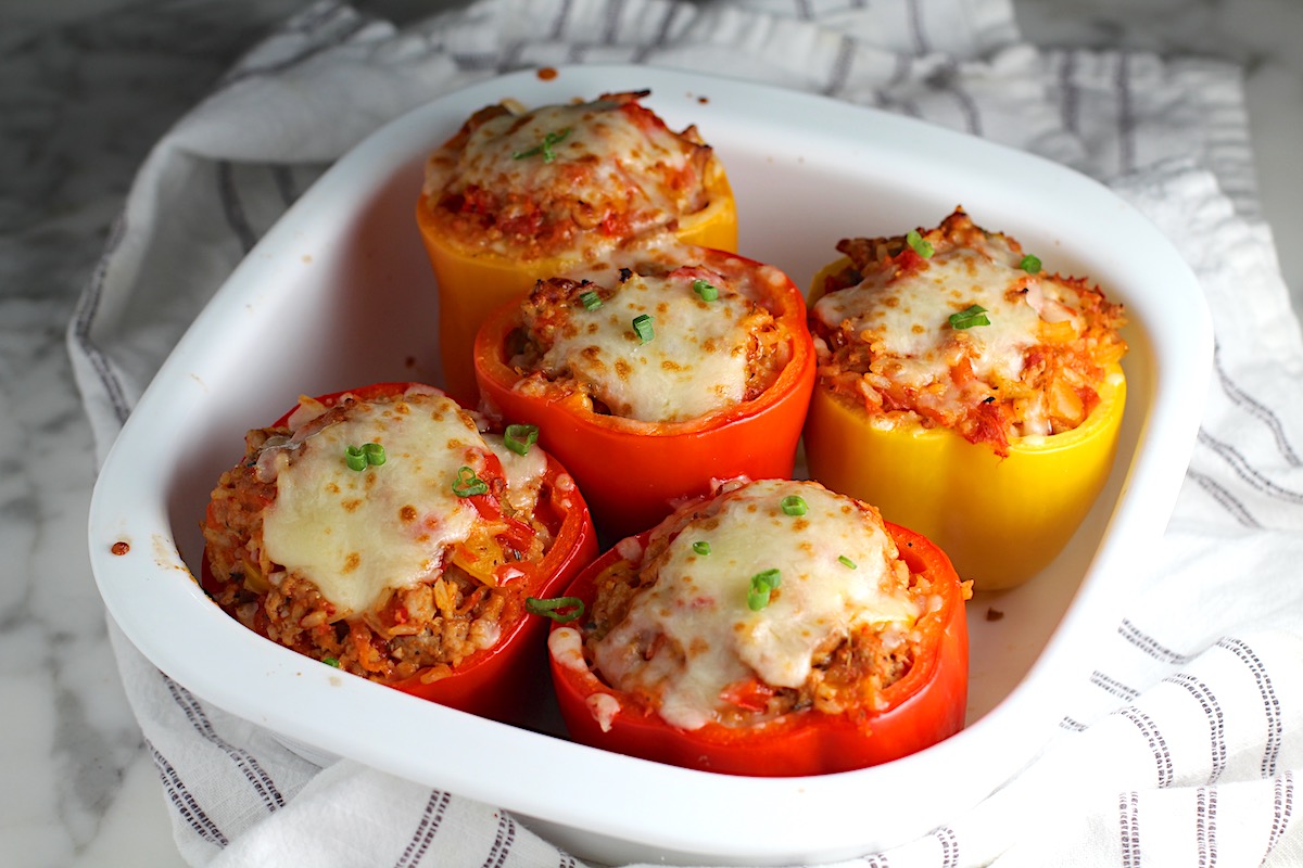 Five Red and Yellow Stuffed peppers with Ground Turkey, rice, and topped with melted cheese and a sprinkle of sliced scallions. The peppers are in the large square casserole dish on a blue and white striped towel on the counter.