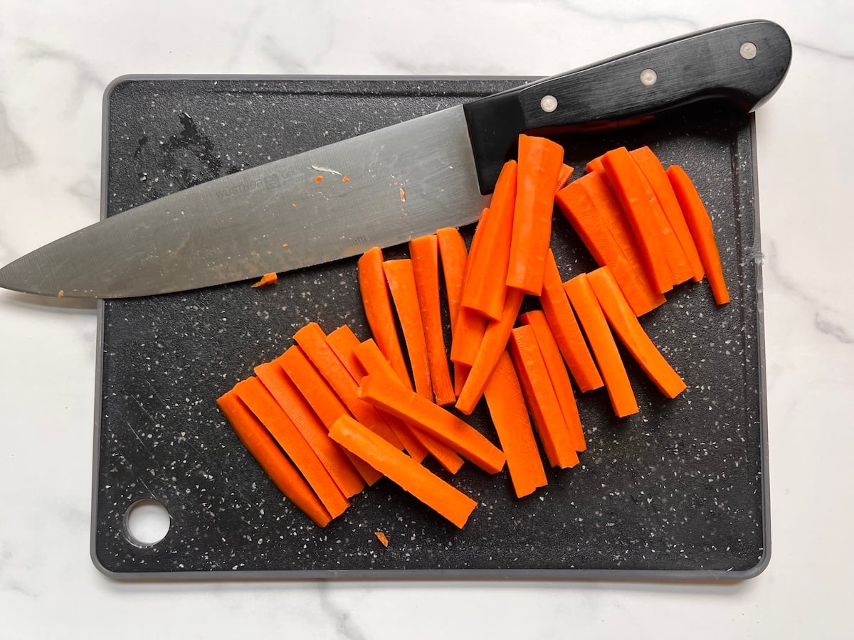 Raw carrots cut into sticks on a black cutting board with chefs knife for this Chicken Stir Fry Recipe.