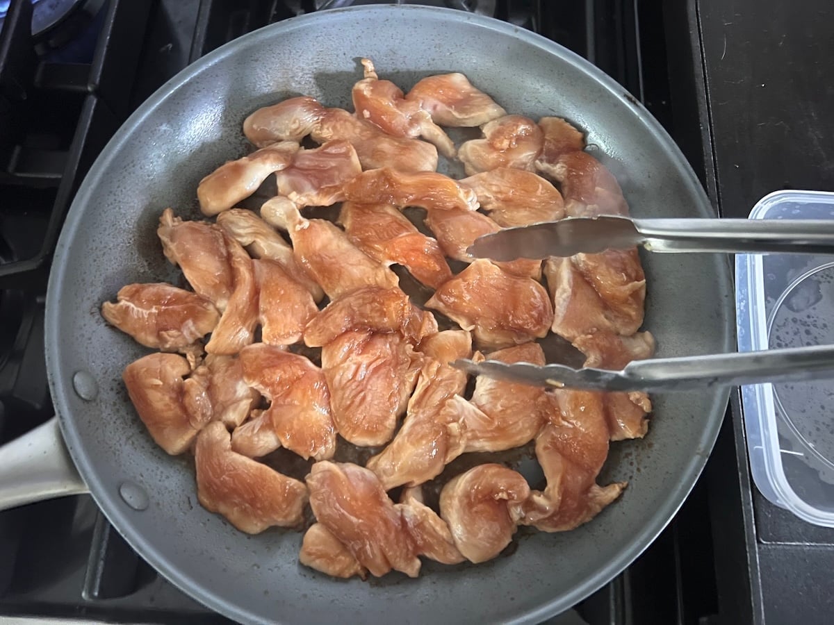 Raw marinated chicken strips cooking in a skillet on stove with tongs about to start flipping pieces over for this Chicken Stir Fry Recipe.