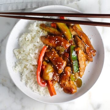 Chicken Stir Fry over white rice on a plate with chopsticks. It has sauce, sliced zucchini, red pepper, celery, onions, carrots and sesame seeds on top. The plate is sitting on a marbled counter with a blue and white striped towel.