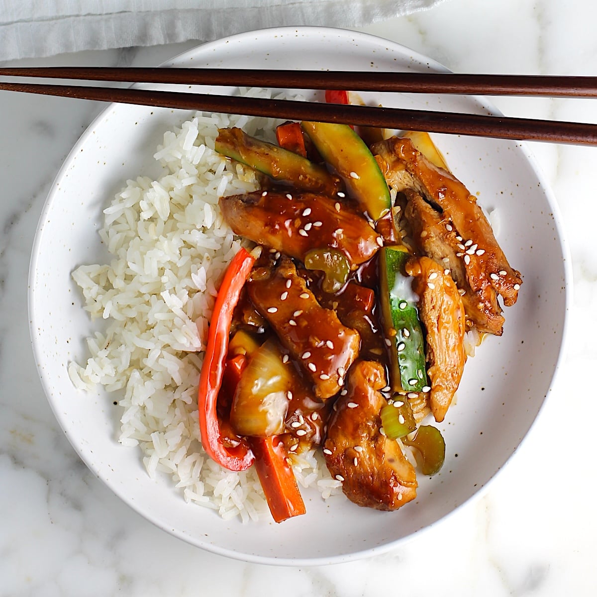 Chicken Stir Fry over white rice on a plate with chopsticks. It has sauce, sliced zucchini, red pepper, celery, onions, carrots and sesame seeds on top. The plate is sitting on a marbled counter with a blue and white striped towel.