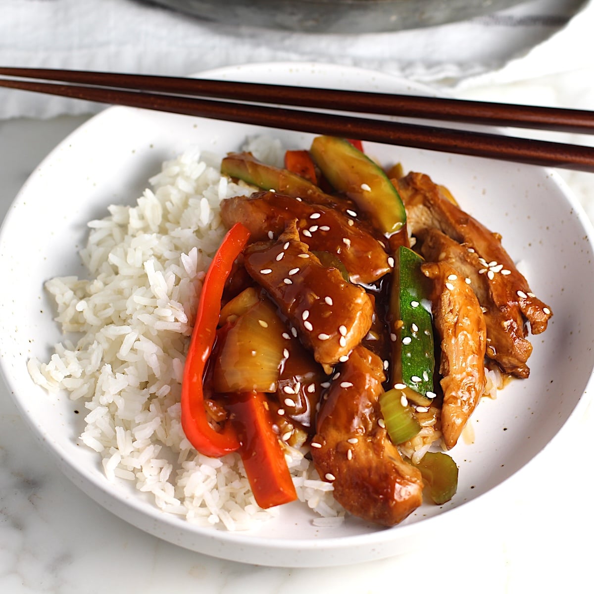 Chicken Stir Fry over white rice on a plate with chopsticks. It has sauce, sliced zucchini, red pepper, celery, onions, carrots and sesame seeds on top. The plate is sitting on a marbled counter with a blue and white striped towel.