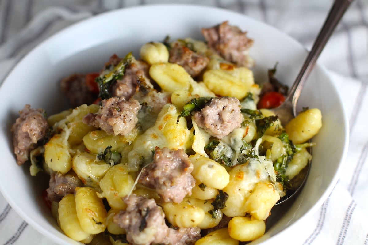 Sheet Pan Gnocchi, Italian Sausage, Broccoli Rabe, and Cherry Tomatoes golden brown with melted mozzarella on top in a serving bowl with spoon.