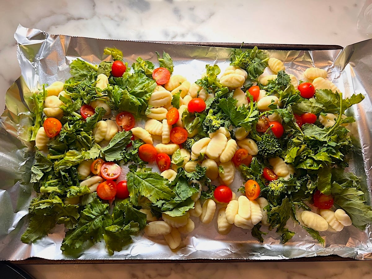 Uncooked Gnocchi, Chopped raw broccoli rabe, and halved cherry tomatoes spread around an aluminum foil lined sheet pan before roasting.