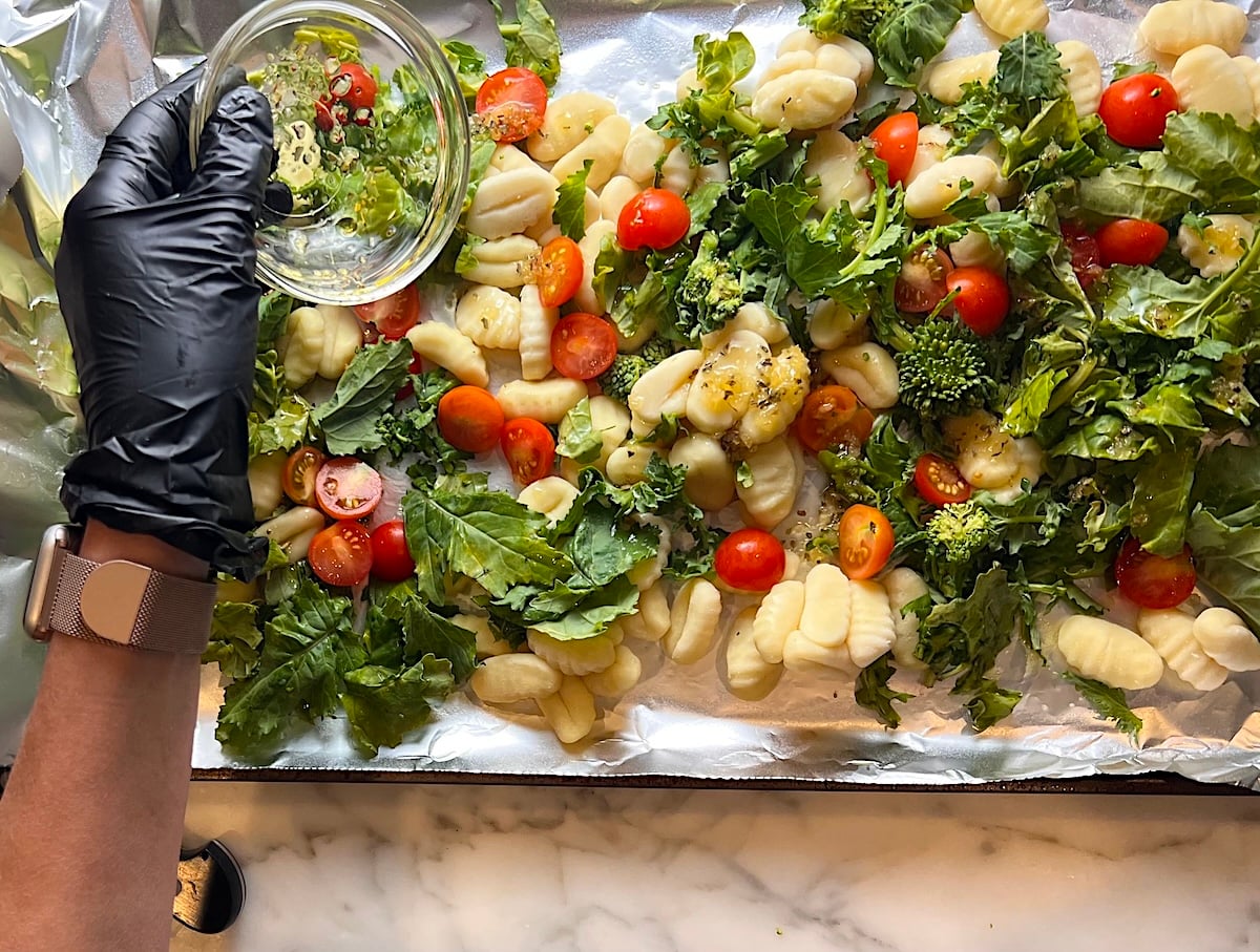 Hand adding olive oil and seasoning to uncooked Gnocchi, Chopped raw broccoli rabe, and halved cherry tomatoes spread around an aluminum foil lined sheet pan before roasting.