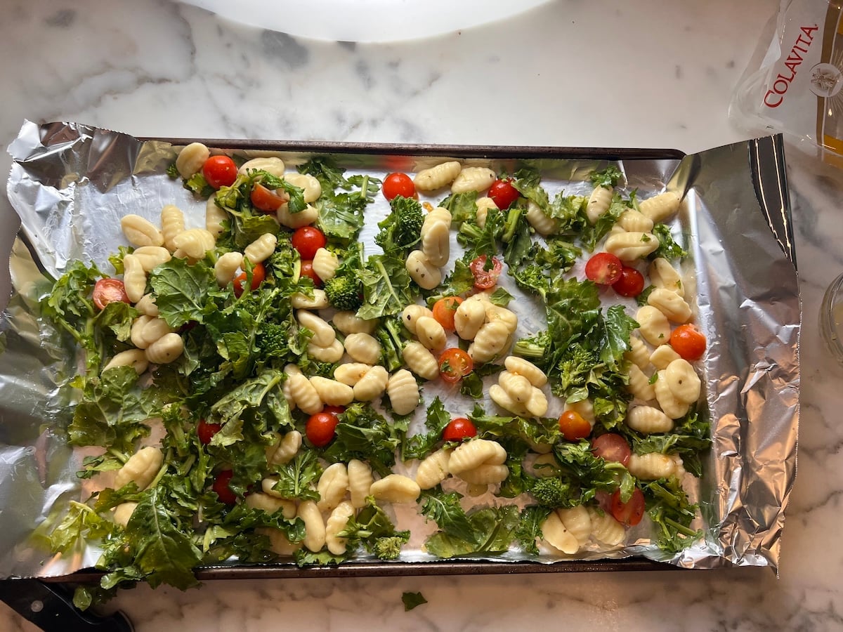 Olive oil, garlic, and seasoning rubbed all over uncooked Gnocchi, Chopped raw broccoli rabe, and halved cherry tomatoes spread around an aluminum foil lined sheet pan before roasting for the recipe, Sheet Pan Gnocchi.