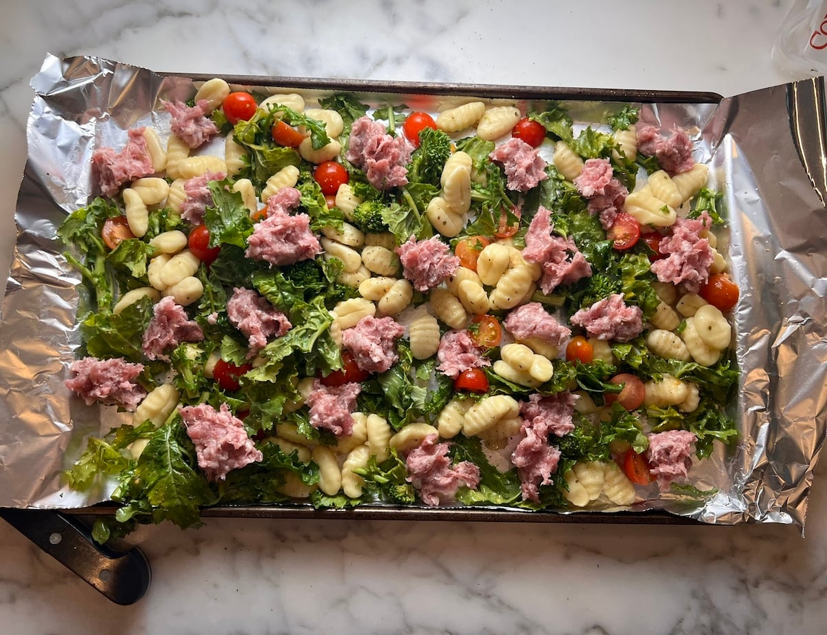 Raw Italian sausage pieces spread around uncooked Gnocchi, Chopped raw broccoli rabe, and halved cherry tomatoes spread around an aluminum foil lined sheet pan before roasting for the recipe, Sheet Pan Gnocchi.