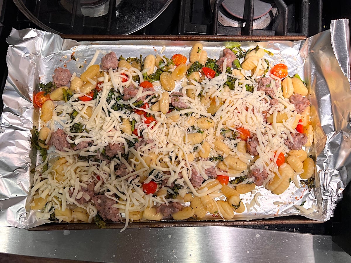 Shredded Mozzarella add on top of cooked Italian sausage pieces, Gnocchi, Chopped raw broccoli rabe, and halved cherry tomatoes on an aluminum foil lined sheet pan for the recipe, Sheet Pan Gnocchi.