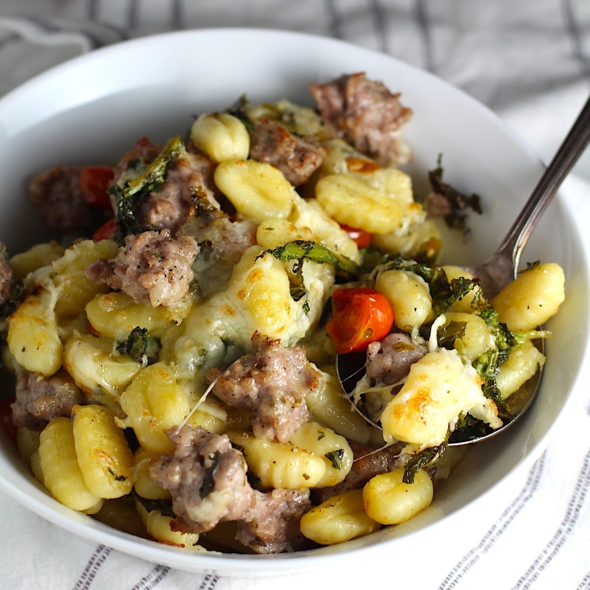 Sheet Pan Gnocchi, Italian Sausage, Broccoli Rabe, and Cherry Tomatoes golden brown with melted mozzarella on top in a serving bowl with spoon.