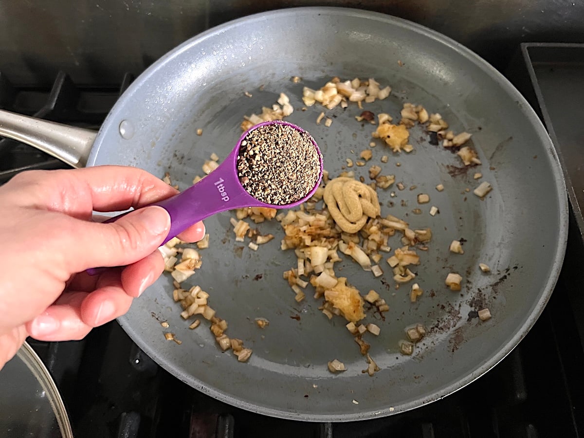 Hand holding a tablespoon of fresh cracked black pepper over a pan with dijon mustard, garlic, and finely diced shallots cooking in the pan after the chicken is transferred out for this Chicken au Poivre recipe.