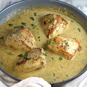 4 pieces of chicken breast seared and cooking in a creamy brown sauce in a pan with seared side-up and browned bits on the bottom of pan still on the stove for this Chicken au Poivre recipe.