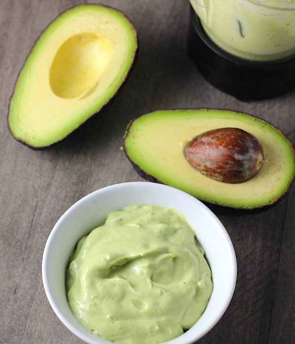 Easy Avocado Crema Recipe in a bowl on table with a cut open avocado with pit and blender in back. It goes on everything! It's a creamy, tangy, rich and delicious sauce that's made in just minutes in the blender!
